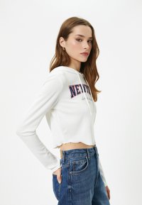 Witte geribde crop sweatshirt met capuchon, voorzien van blauwe en rode "NEW YORK" tekst. Gekoppeld aan hoge taille blauwe denim jeans.