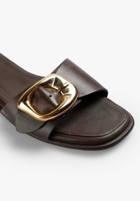 Brune lædersandale med bred rem og et stort, guld rektangulært spænde som detalje. Glat overflade med stikninger.