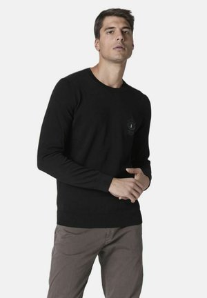 Maglione nero a maniche lunghe con scollo rotondo e polsini a coste. Presenta un piccolo logo testurizzato sul petto. Abbinato a pantaloni grigi chiari.