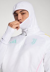 Fehér kapucnis sportdzseki fél cipzárral, mentazöld és sárga dizájnelemekkel, sima tapintású, karcsúsított szabású.