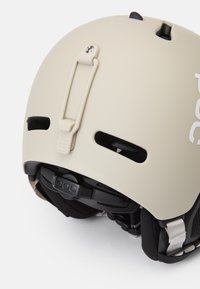 Hellbeige Helm mit Belüftungsöffnungen, verstellbaren Riemen und einem schwarzen Einstellrad, mit einem auffälligen weißen Logo an der Seite.