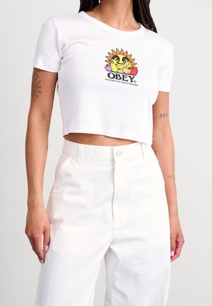 Femme portant un t-shirt blanc court avec un graphique coloré de soleil et de fruits et le texte « OBEY », associé à un pantalon blanc taille haute.
