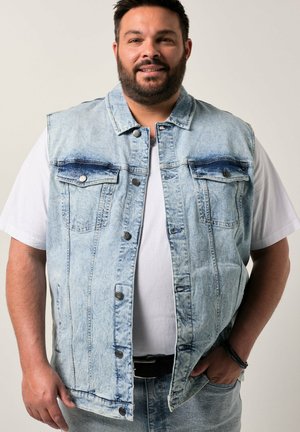 Mand med skæg iført lyseblåt denimvest over hvid t-shirt, stående med den ene hånd i lommen mod en ensfarvet baggrund.