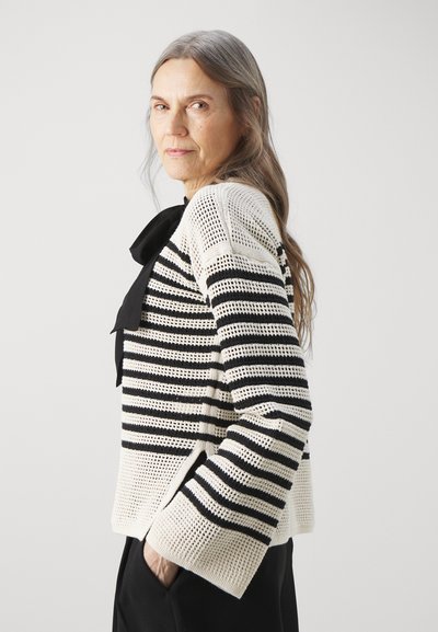 Pull en tricot avec des rayures horizontales noires et blanches, présentant un décolleté large, des manches longues et un tissu texturé. Accent nœud à l'avant.