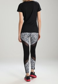 Woman seen from behind wearing black short-sleeve top, patterned leggings with black mesh inserts, and black sneakers with red soles.