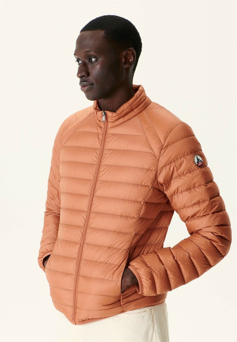 Giacca imbottita di colore arancione bruciato, con colletto alto, chiusura con zip e tasche laterali. Strisce texturizzate corrono verticalmente lungo le maniche.