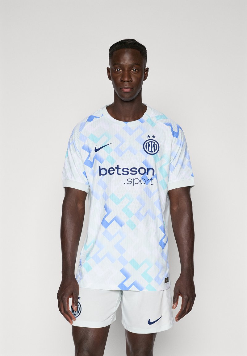 Fehér sportmez kék geometrikus mintákkal, Nike logóval és "betsson.sport" felirattal. Fehér sportnadrággal párosítva. A textúra textúrált hatású.