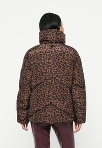 Cilvēks valkā brūnu un melnu leopardu rakstu pūkainu virsjacku ar augstu apkakli, redzams no mugurpuses uz balta fona.