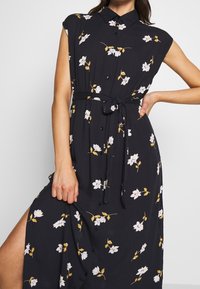 Schwarzes Blumenkleid mit kurzen Ärmeln, Kragen, durchgehender Knopfleiste und gebundenem Taillenband. Mit weißen und gelben Blumenmustern über das gesamte Kleid.