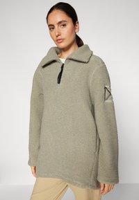 Pullover in pile di colore verde oliva con colletto con zip, fessure laterali e una toppa triangolare sulla manica sinistra. Materiale testurizzato e accogliente.
