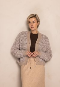 Hellbeige kurz geschnittener Cardigan aus strukturiertem, flauschigem Stoff, mit gerippten Bündchen und Taillenbund, braunen Knöpfen, über einem dunkelbraunen Top getragen.