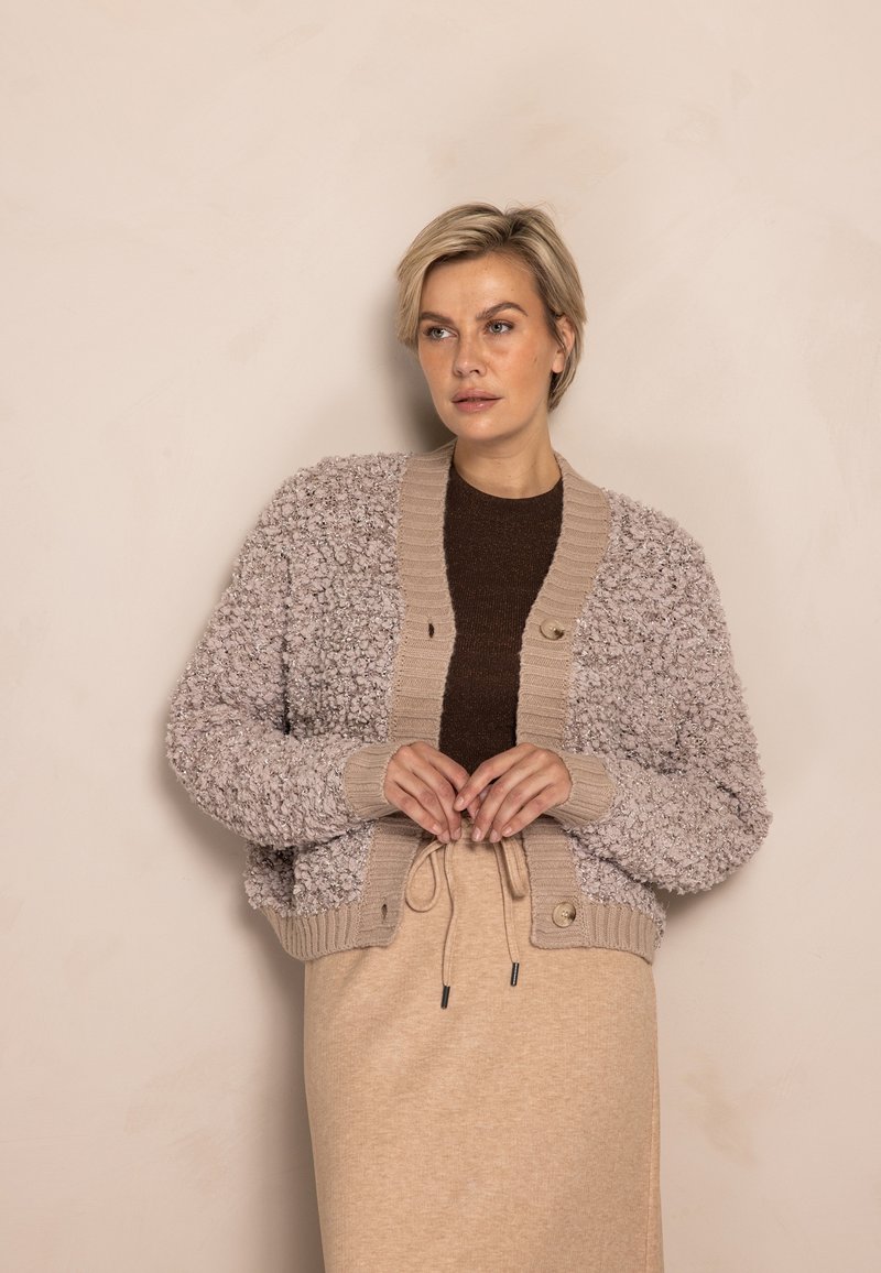 Hellbeige kurz geschnittener Cardigan aus strukturiertem, flauschigem Stoff, mit gerippten Bündchen und Taillenbund, braunen Knöpfen, über einem dunkelbraunen Top getragen.