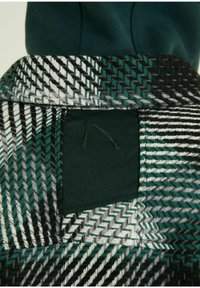 Närbild av grönt, vitt, svart och grått vävt ulltyg med fiskbensmönster och en vikt krage över ett mörkgrönt plagg.