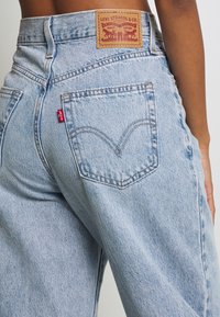 Záběr zblízka na osobu v světle modrých džínách od Levi's, ukazující zadní kapsu a koženou značkovou nášivku Levi's na pase.