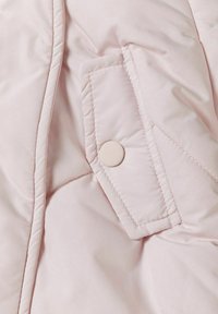 Lichtroze pufferjacksweef met diagonaal quiltpatroon. Voorzien van een patchzak die is beveiligd met een ronde drukknoop. Gladde textuur.