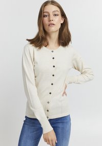Cardigan tricotat bej, cu guler rotund, nasturi negri pe față și mâneci lungi cu un design ajustat.