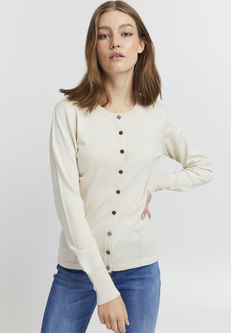 Cardigan en tricot beige avec un col rond, des boutons noirs sur le devant et des manches longues avec une coupe ajustée.