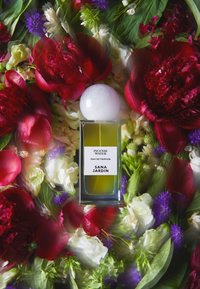 Bouteille de parfum en verre avec un capuchon rond blanc, liquide vert, et une étiquette présentant "Eau d'Encens" et "Sana Jardin", entourée de fleurs colorées.