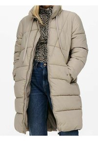 Lang beigefarvet quiltet pufferjakke med hætte, båret over en leopardmønstret turtleneck og blå højtaljede jeans.