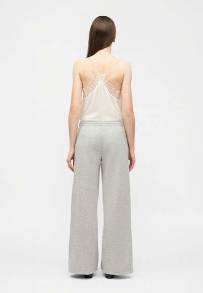 Femme aux longs cheveux bruns portant un haut blanc transparent sans manches avec des volants et un pantalon large ample gris clair, debout de dos.