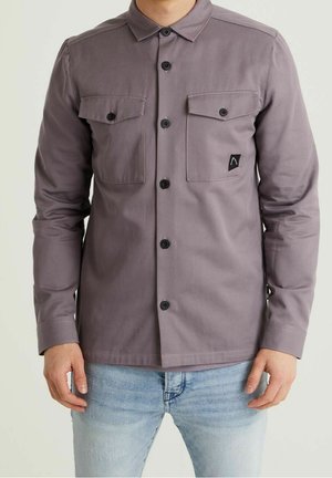 Homme portant une chemise grise à manches longues boutonnée avec deux poches poitrine et un jean bleu clair, debout devant un fond uni.