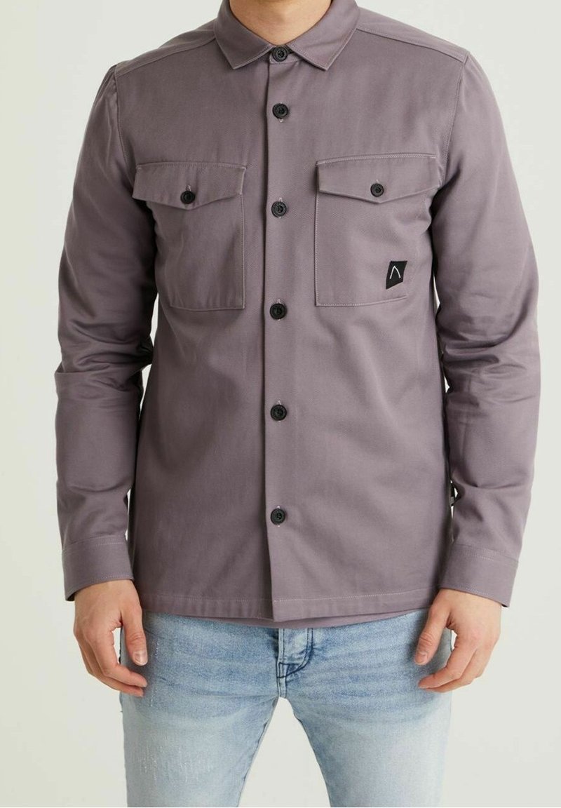 Homme portant une chemise grise à manches longues boutonnée avec deux poches poitrine et un jean bleu clair, debout devant un fond uni.