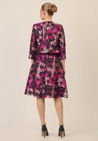 Conjunto de vestido floral y chaqueta en tela oscura con grandes flores rosas y moradas. Falda hasta la rodilla con cintura ajustada, acompañada de tacones altos.