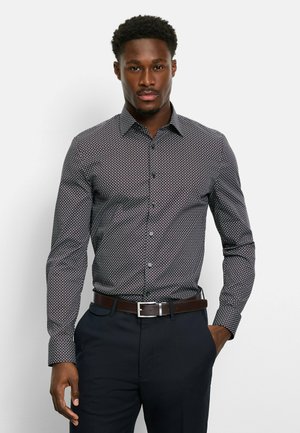 Marineblaue Button-Up-Hemd mit einem geometrischen Muster in Weiß und Rot. Lange Ärmel, klassischer Kragen und kombiniert mit dunklen Hosen und einem braunen Gürtel.