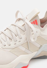 Light beige athletic shoe with textured fabric upper, narrow white laces, and orange accents. Features a cushioned sole and branding details.