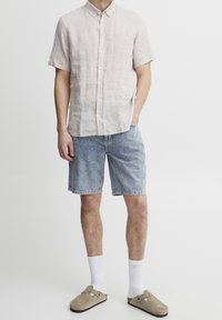 Light beige linen short-sleeve shirt with button-front, blue denim shorts, white socks, and beige slip-on shoes with two holes.