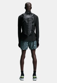 Black backpack with a sleek, streamlined design featuring adjustable straps and a top handle. Worn by a person in black long sleeves and patterned shorts.