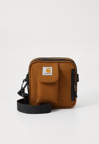 Pruun kangast rihmakott esiflapiga taskuga, musta tõmbelukuga, reguleeritava rihmaga ning Carhartti logo patchiga oranžis ja valges.