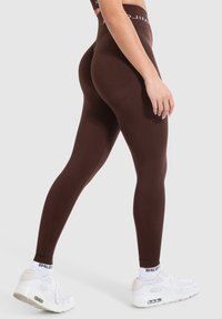 Leggings marroni a vita alta realizzati in materiale elasticizzato, con una texture liscia e una fascia con logo bianco in vita. Indossati con sneakers bianche.