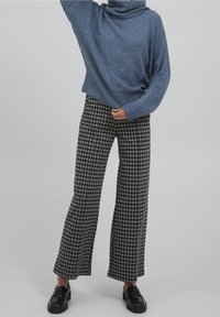 Person, der einen blauen Rollkragenpullover, schwarze und weiße karierten weit geschnittenen Hosen und schwarze Slipper trägt, steht vor einem schlichten Hintergrund.