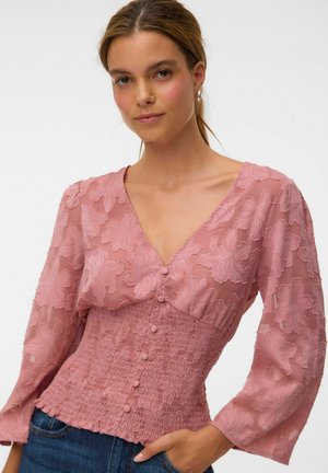 Femme portant un chemisier rose à fleurs texturé avec des boutons devant et un jean bleu, posant avec une main dans la poche.