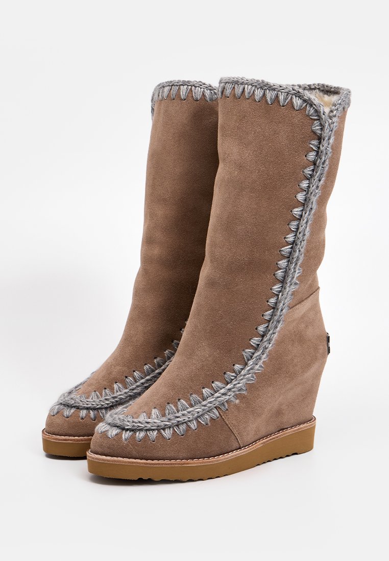 Botas de cuña de gamuza beige con un caño alto, que presentan un intrincado bordado gris a lo largo de las costuras y un suave forro interior.