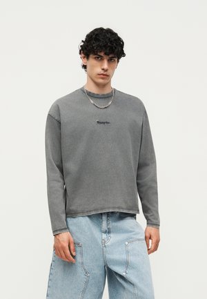 Νεαρός με σγουρά σκούρα μαλλιά, φοράει ξεθωριασμένο γκρι μακρυμάνικο πουκάμισο και ανοιχτό μπλε φαρδιά τζιν, με φόντο απλό.