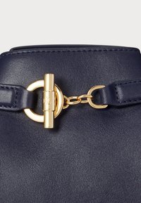 Szczegół damskiej torebki skórzanej w kolorze granatowym z złotym okrągłym akcentem oraz prostokątnym zapięciem, o fakturowym wykończeniu.