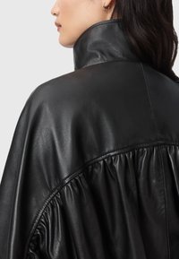 Žena v černé kožené bundě s vysokým límcem a na zadní straně s nařasením, doplněná o geometrický stříbrný náušnici.