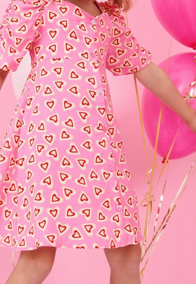 Enfant portant une robe rose avec des motifs de cœurs rouges et blancs, tenant des ballons roses avec des rubans dorés devant un fond rose.