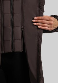 Giacca imbottita marrone con texture trapuntata, con doppie zip frontali e una finitura liscia e opaca. In primo piano il dettaglio della zip.