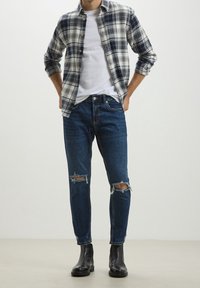 Man wearing rolled-up plaid shirt over white t-shirt, ripped dark blue jeans, and black leather ankle boots standing with hands in pockets.