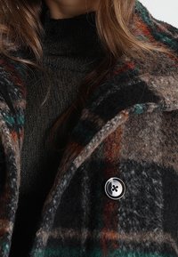 Uldfrakke med tern i en blanding af brun, sort og grøn med røde accenter. Indeholder en rund knapdetalje og en tekstureret krave.