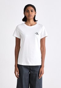 White cotton t-shirt with round neck and short sleeves. Features a small black logo on the chest. Paired with dark denim wide-leg pants.