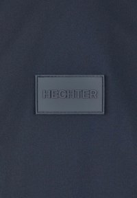 Navyfarbener Stoff mit glatter Textur zeigt ein rechteckiges Gummipatch, das in der Mitte mit "HECHTER" geprägt ist, ohne weitere sichtbare Details.