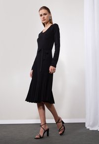 Schwarzes, langärmliges Kleid mit tailliertem Oberteil, gefälteltem Rock und gebundenem Taillendetail. Begleitet von schwarzen Riemchensandalen. Glatte Textur.