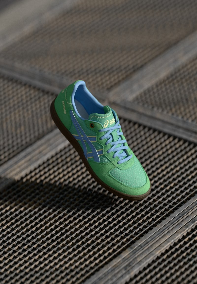 Zaļas sporta kurpes ar teksturētu virsmu, zilām akcentiem un gaiši zilām auklām. Iekļauj gludu purngalu un brūnu gumijas zoli.