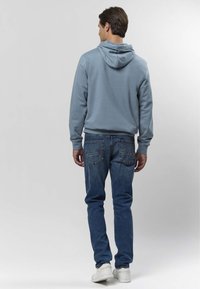 Svetlo modra pulover s kapuco in kenguru žepom, kombinirana z modrimi denim kavbojkami in belimi supergami, gledano od zadaj.