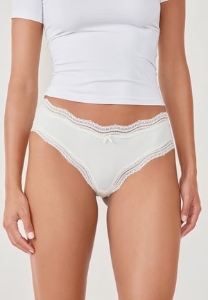 Mutande bianche con pizzo e un delicato fiocco davanti, realizzate in un tessuto morbido e con bordi ondulati, messe in risalto da una pelle liscia.