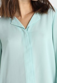 Blusa de color azul claro, con aspecto de seda, con cuello acentuado y tapeta frontal. Textura suave y corte holgado con mangas largas.